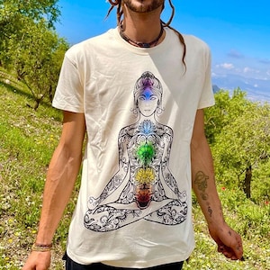 Könnte beinhalten: Ein weißes T-Shirt mit einer schwarz-weißen Illustration einer Person in einer Yoga-Pose. Die Illustration enthält sieben Chakren, jedes mit einer anderen Farbe. Die Chakren befinden sich am Fuß der Wirbelsäule, am Kreuzbein, am Solarplexus, am Herzen, am Hals, am dritten Auge und an der Krone.