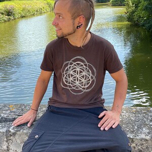 May include: A person wearing a brown t-shirt with a white geometric design and black pants. The person is sitting on a stone wall by a pond.