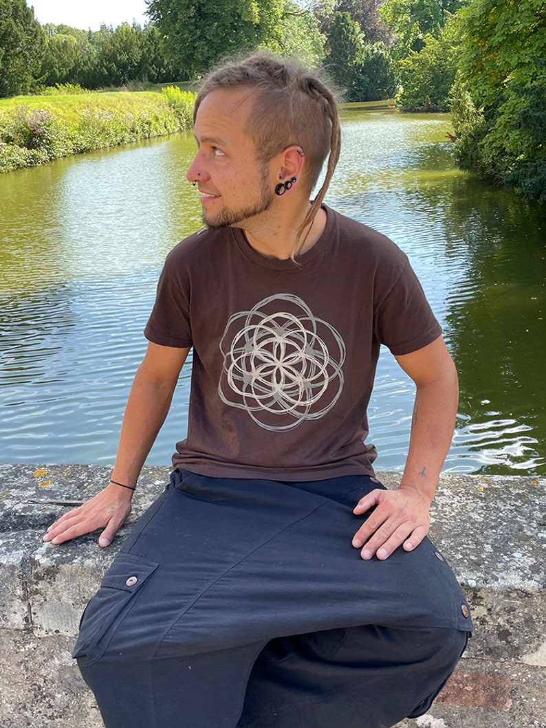 May include: A person wearing a brown t-shirt with a white geometric design and black pants. The person is sitting on a stone wall by a pond.