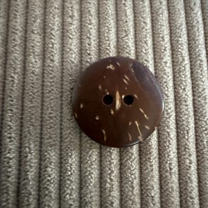 Könnte beinhalten: Ein runder, dunkelbrauner Knopf mit zwei Löchern und hellen Sprenkeln. Der Knopf besteht aus einem natürlichen Material und hat eine glatte, leicht glänzende Oberfläche. Er liegt vor einem strukturierten, hellbraunen Hintergrund.
