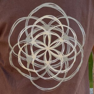 May include: A brown t-shirt with a white geometric design printed on the front. The design is a circle with overlapping circles inside, creating a flower-like pattern.