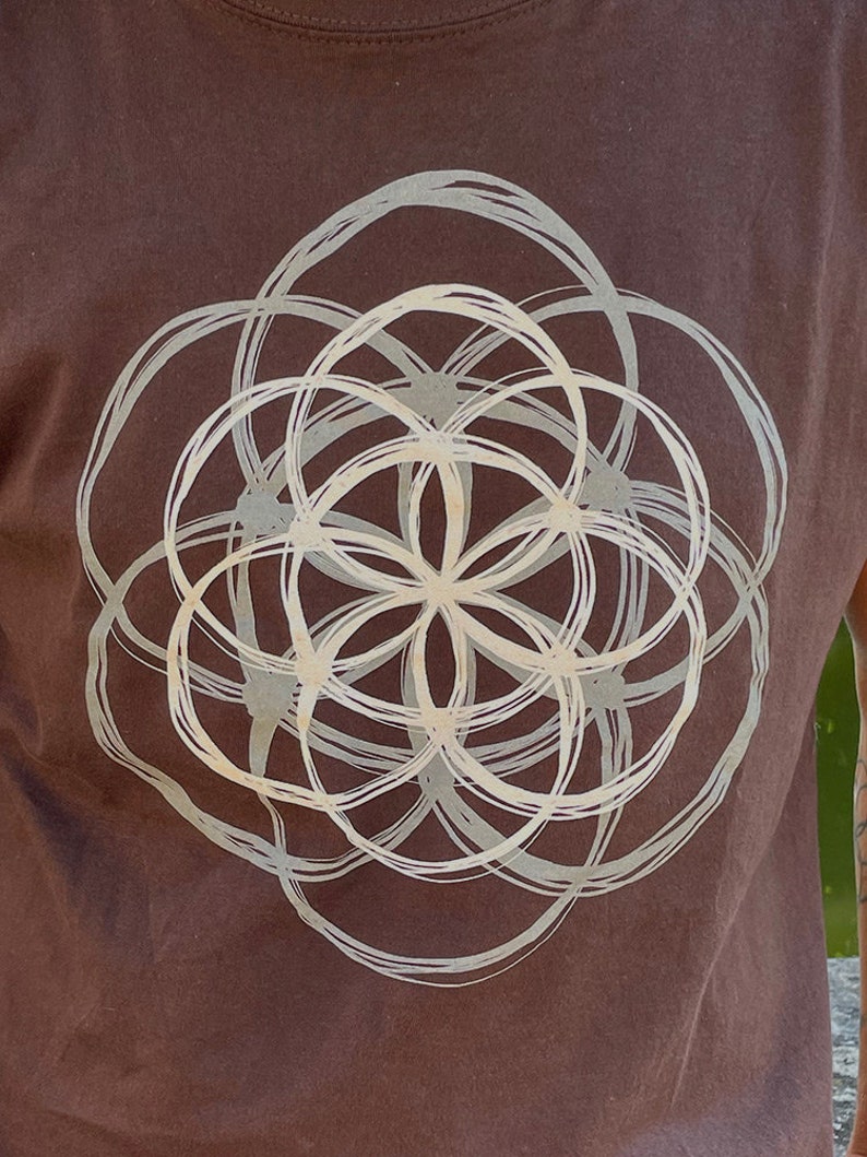 May include: A brown t-shirt with a white geometric design printed on the front. The design is a circle with overlapping circles inside, creating a flower-like pattern.
