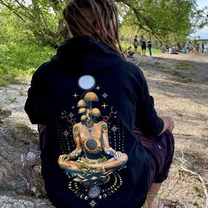 Könnte beinhalten: Schwarzer Kapuzenpullover mit einem psychedelischen Motiv einer meditierenden Figur mit Pilzen, die aus ihrem Kopf wachsen. Das Motiv enthält einen Mond, Sterne und geometrische Muster.