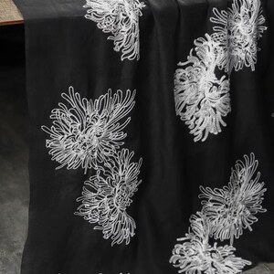 May include: A black fabric runner featuring an embroidered pattern of white chrysanthemum flowers. The floral design is scattered across the surface, showcasing intricate thread work that creates a contrast against the dark background.
