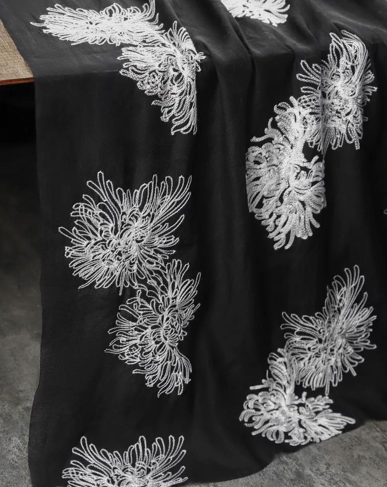 May include: A black fabric runner featuring an embroidered pattern of white chrysanthemum flowers. The floral design is scattered across the surface, showcasing intricate thread work that creates a contrast against the dark background.