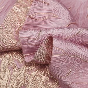 Pode incluir: Tecido rosa com um padrão tecido de linhas metálicas douradas. O tecido é texturizado e tem um brilho sutil.
