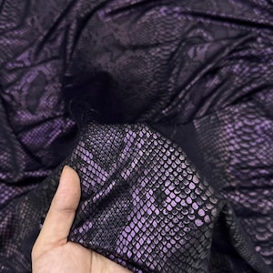 Könnte beinhalten: Nahaufnahme eines dunkelvioletten Stoffes mit Schlangenmuster. Der Stoff hat eine glänzende, strukturierte Oberfläche mit einem schuppenartigen Design. Das Material ist gerafft und drapiert, wodurch die Textur- und Farbvariationen sichtbar werden.