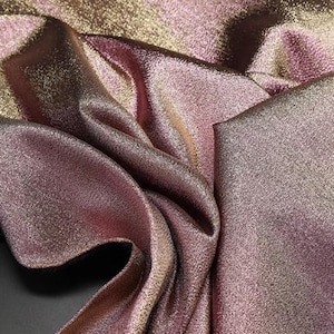 May include: Close-up of a metallic pink and gold fabric with a subtle shimmer. The fabric is folded and draped, showcasing its texture and sheen.