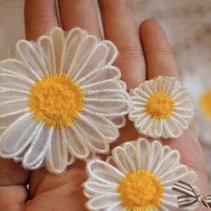 Puede incluir: Tres aplicaciones de tela en forma de margarita blancas con centros amarillos. Las aplicaciones están dispuestas en orden decreciente de tamaño en una mano.