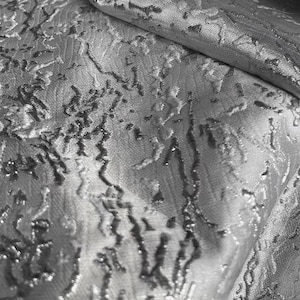 Może przedstawiać: Zbliżenie srebrnej tkaniny z fakturowanym, tłoczonym wzorem. Tkanina ma subtelny połysk i lekko pomarszczony wygląd.