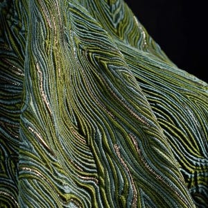 Puede incluir: Primer plano de una tela texturizada con un patrón fluido y orgánico. La tela presenta tonos de verde y turquesa con acentos metálicos dorados. El diseño se asemeja a las líneas de un mapa topográfico o a la veta de la madera.