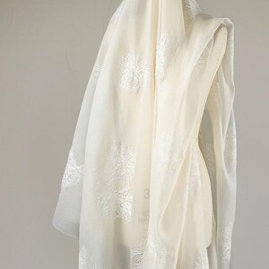 May include: A sheer, off-white fabric drape featuring an intricate, raised white floral embroidery pattern throughout. The lightweight material is layered over a dress form, showcasing the delicate texture and semi-transparent quality of the textile.