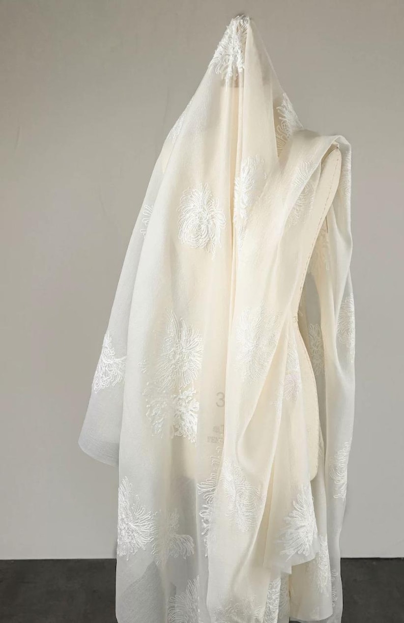 May include: A sheer, off-white fabric drape featuring an intricate, raised white floral embroidery pattern throughout. The lightweight material is layered over a dress form, showcasing the delicate texture and semi-transparent quality of the textile.