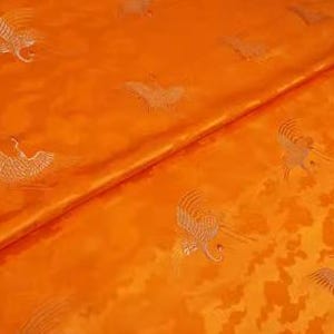 May include: Orange fabric with a repeating pattern of white embroidered cranes. The fabric has a subtle sheen and a textured surface.