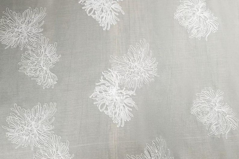 May include: A sheer light gray fabric featuring an embroidered pattern of white chrysanthemum flowers. The delicate floral design is repeated across the surface, showcasing intricate line work that defines each petal of the blossoms.