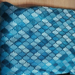 Op de afbeelding: Turquoise stof met een wit en turquoise geometrisch patroon van overlappende waaiervormige vormen.