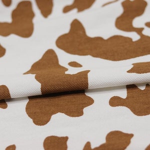 K&ouml;nnte beinhalten: Wei&szlig;er Stoff mit einem braunen Kuhmuster. Der Stoff ist in der Mitte gefaltet und zeigt die Textur des Materials.