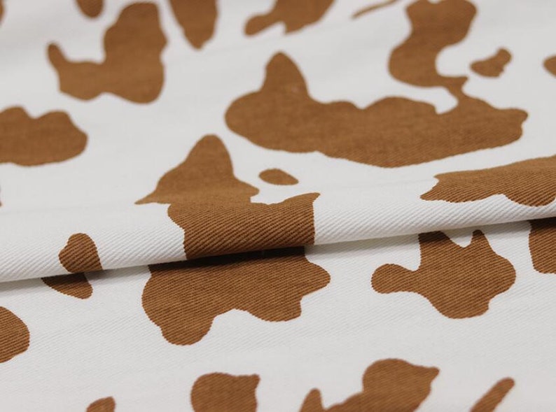 K&ouml;nnte beinhalten: Wei&szlig;er Stoff mit einem braunen Kuhmuster. Der Stoff ist in der Mitte gefaltet und zeigt die Textur des Materials.