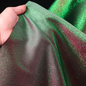 May include: A piece of iridescent fabric featuring a color-shifting effect that transitions between vibrant emerald green and deep magenta. The material has a fine, textured weave with a metallic sheen that catches the light across its surface.