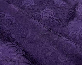 Può includere: Primo piano di un tessuto viola scuro con un motivo floreale. Il tessuto presenta rose e foglie in rilievo, creando una superficie strutturata. Il design si ripete sul materiale, mostrando uno stile classico ed elegante.