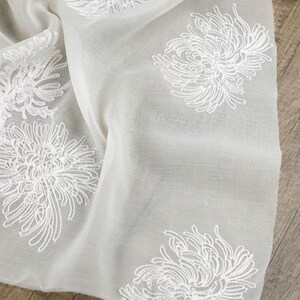 May include: A sheer, off-white fabric scarf featuring an embroidered pattern of white chrysanthemum flowers. The lightweight material has a soft, semi-transparent texture with delicate white thread work detailing the floral motifs across the surface.