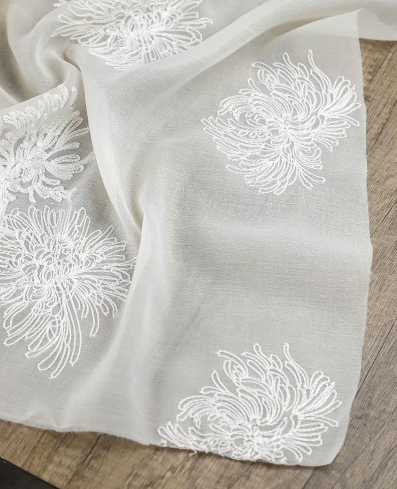 May include: A sheer, off-white fabric scarf featuring an embroidered pattern of white chrysanthemum flowers. The lightweight material has a soft, semi-transparent texture with delicate white thread work detailing the floral motifs across the surface.