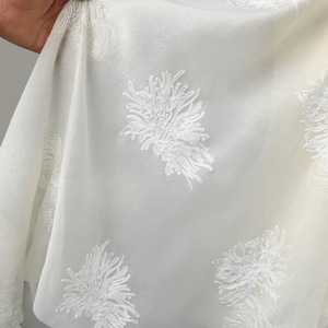 May include: A sheer white fabric featuring an embroidered pattern of raised, textured white floral motifs. The material is lightweight and semi-transparent, with the intricate threadwork creating a distinct, three-dimensional appearance.