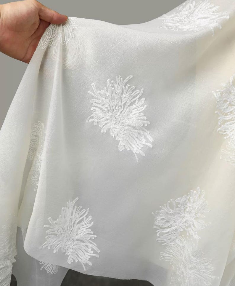 May include: A sheer white fabric featuring an embroidered pattern of raised, textured white floral motifs. The material is lightweight and semi-transparent, with the intricate threadwork creating a distinct, three-dimensional appearance.