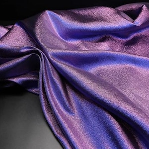 May include: A close-up of shimmering fabric in shades of purple and blue. The material is draped, creating folds and shadows that highlight its texture and sheen. The fabric appears to be a luxurious textile, possibly for apparel or decorative use.