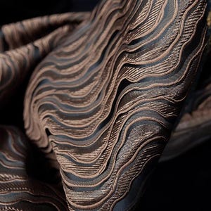 Puede incluir: Primer plano de una tela marrón con un patrón ondulado y texturizado. La tela tiene un brillo sutil y una textura ligeramente en relieve.