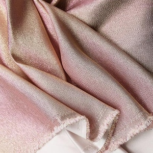 May include: Close-up of a shimmering fabric with a gradient of pink, gold, and bronze hues. The material is draped, revealing a textured surface and a raw, fringed edge. The fabric appears to be lightweight and flowing, with a subtle sheen.