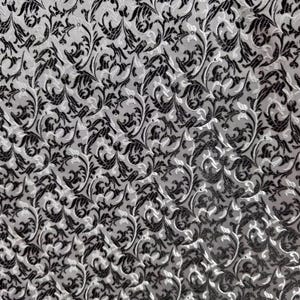 May include: Close-up of a fabric with a repeating pattern of black and white floral designs. The pattern features stylized leaves and swirling lines, creating a textured, elegant appearance. The fabric has a subtle sheen.