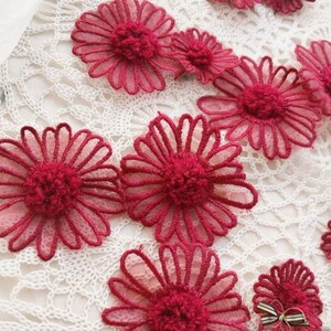 May include: A collection of dark red daisy-shaped fabric appliques. The appliques feature a sheer, petal design with a textured centre. They are arranged on a white, open-weave fabric background, creating a delicate floral theme.