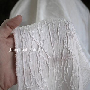 Puede incluir: Tela jacquard blanca con un patrón en relieve de hojas y ramas. La tela es suave y tiene un brillo sutil. "Jacquard Fabric" está escrito en negro en la tela.