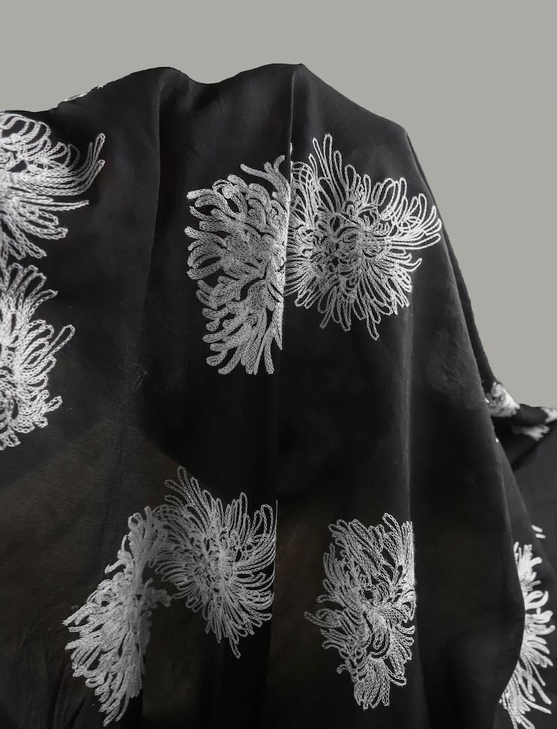 May include: A black fabric textile featuring a repeating pattern of white embroidered chrysanthemum flowers. The material is lightweight and semi-sheer, with the intricate white thread work creating a high-contrast floral design across the surface.