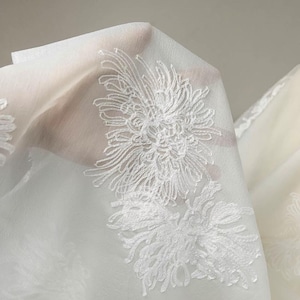 May include: A sheer white fabric featuring intricate white floral embroidery. The delicate material is translucent, showcasing detailed threadwork of chrysanthemum-like blossoms scattered across the surface in a repeating pattern.