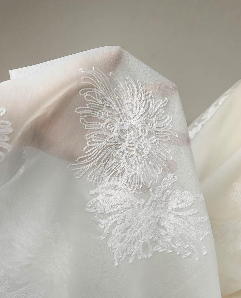 May include: A sheer white fabric featuring intricate white floral embroidery. The delicate material is translucent, showcasing detailed threadwork of chrysanthemum-like blossoms scattered across the surface in a repeating pattern.