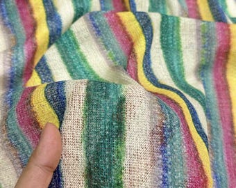 May include: Close-up of a colorful fabric with vertical stripes in shades of yellow, blue, green, and pink. The fabric has a textured appearance and is held in a hand, showing its drape and weave. The background is blurred, emphasizing the textile's pattern.