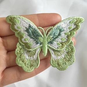 Puede incluir: Un parche de aplique de mariposa bordado en verde y blanco. La mariposa tiene un diseño detallado con un borde festoneado.
