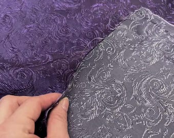 May include: Close-up of two fabric swatches. One is a deep purple with a raised, swirling pattern. The other is a gray fabric with a similar, but lighter, swirling design. The fabrics are likely for upholstery or decorative purposes.