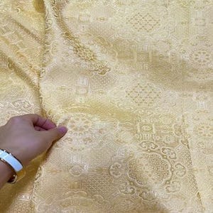 Può includere: Primo piano di un tessuto dorato con un motivo bianco intricato. Il tessuto ha una lucentezza sottile e una sensazione di lusso.