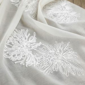May include: A sheer, off-white fabric scarf featuring delicate, white embroidered floral patterns that resemble chrysanthemums. The lightweight material is draped in soft folds, highlighting the intricate threadwork of the floral designs.
