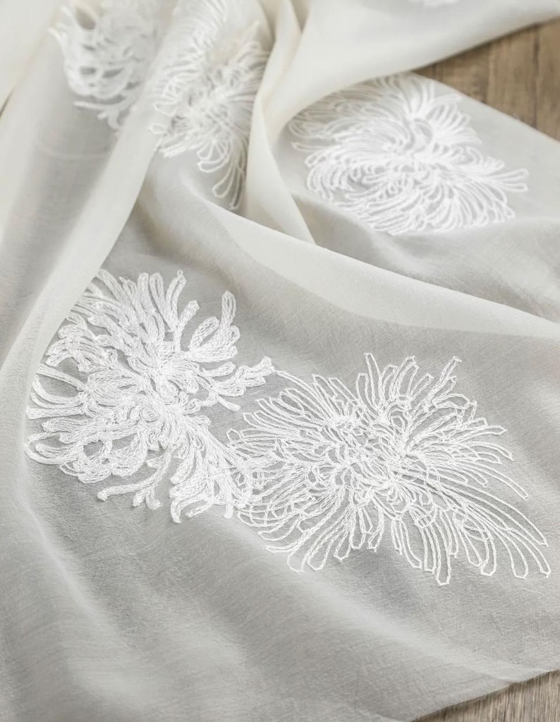 May include: A sheer, off-white fabric scarf featuring delicate, white embroidered floral patterns that resemble chrysanthemums. The lightweight material is draped in soft folds, highlighting the intricate threadwork of the floral designs.