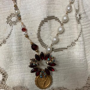 Op de afbeelding: Een vintage-stijl ketting met imitatieparels, rode en heldere facetkralen en goudkleurige metalen accenten. Het middelpunt is een hanger met een gouden munt en een cluster van rode en iriserende kristallen.