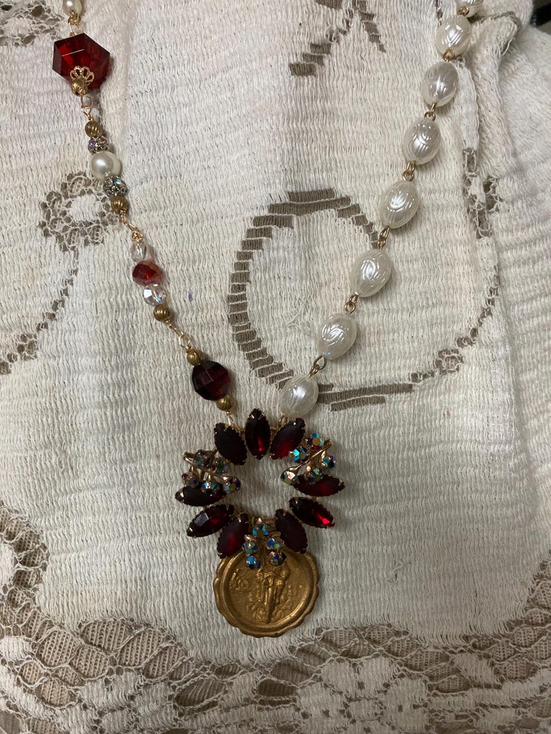 Op de afbeelding: Een vintage-stijl ketting met imitatieparels, rode en heldere facetkralen en goudkleurige metalen accenten. Het middelpunt is een hanger met een gouden munt en een cluster van rode en iriserende kristallen.