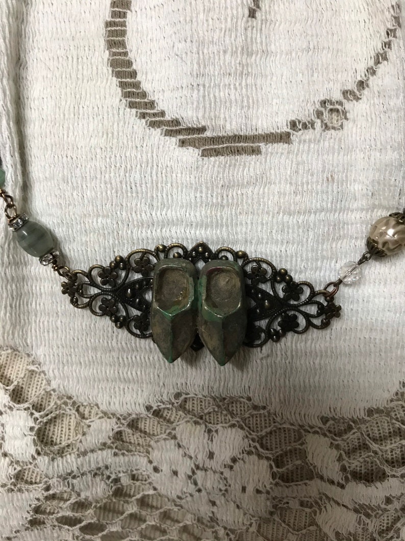 May include: A necklace with a bronze filigree pendant featuring two miniature green shoes. The necklace also has green and clear beads.