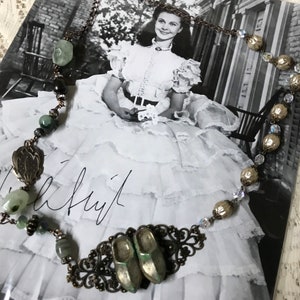 May include: A vintage-inspired necklace with green and brown beads, a metal filigree design, and two small green ceramic shoes. The necklace is displayed on a black and white photo of a woman in a dress.