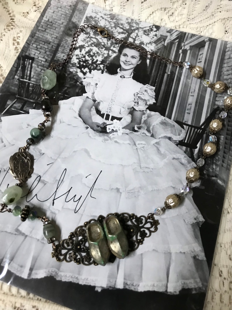 May include: A vintage-inspired necklace with green and brown beads, a metal filigree design, and two small green ceramic shoes. The necklace is displayed on a black and white photo of a woman in a dress.