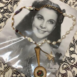 May include: A vintage-style necklace featuring a gold-toned chain, faux pearls, and decorative beads. A gold-toned spoon pendant with a floral embroidered design and a small fairy charm hangs from the necklace. The necklace is displayed on a black and white photograph.