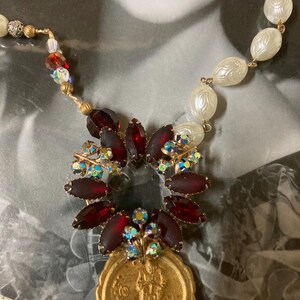 Op de afbeelding: Vintage ketting met een gouden hanger met een figuur, omringd door donkerrode en iriserende kristallen. De ketting bevat witte, getextureerde kralen en kleinere, kleurrijke kralen. Het ontwerp is geplaatst tegen een vintage foto.